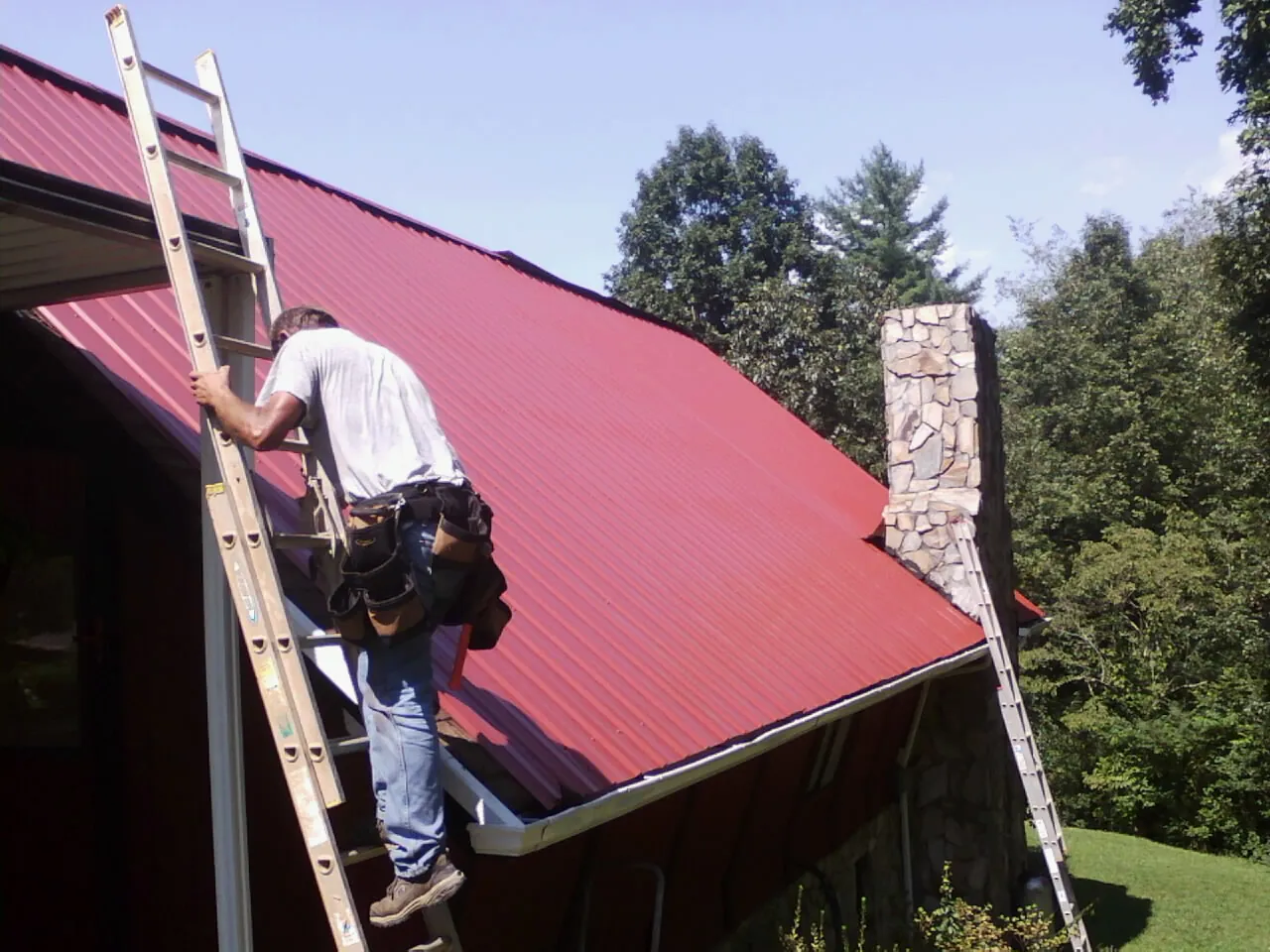 Roofer on a ladder beside a red metal roof and stone chimney — Soffit & Fascia Repair by Roofing Brothers in Sparta